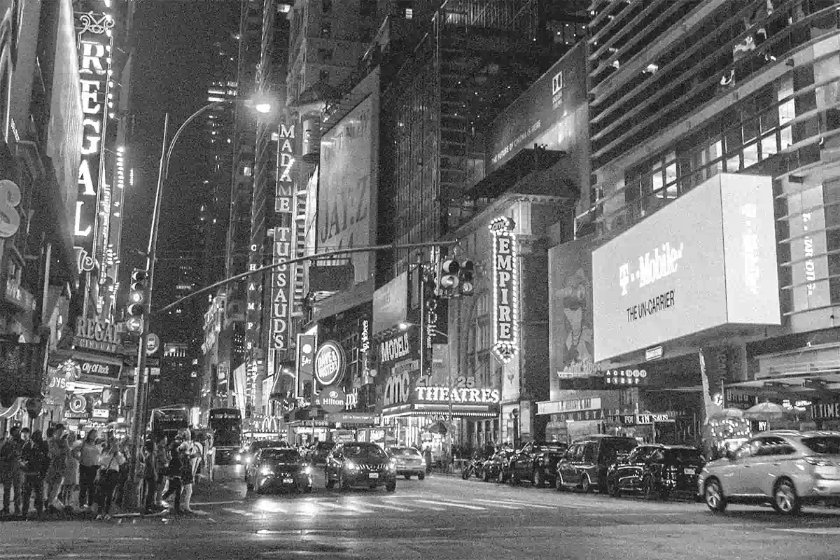 Ein Schwarz-weiß-Foto vom Times Square in New York. Es ist dunkel und sehr viele Menschen und Autos sind unterwegs. Überall leuchtet Reklame-Werbung.